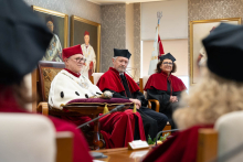 Trzy osoby osoby siedzą na fotelach. Przed nimi leżą insygnia na poduszce. W tle widoczne są portrety i flaga.