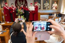 Kilka osób stoi przy podwyższeniu i pozuje do zdjęcia. Na pierwszym planie widoczne są ręce trzymające telefony.