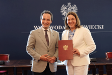 Mężczyzna po lewej i kobieta po prawej stoją przed tłem z napisem „Wojewoda Mazowiecki” i godłem Polski. Osoby wspólnie trzymają zamkniętą czerwoną teczkę z godłem.