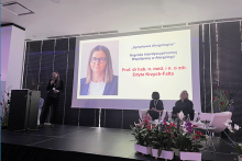 Ekran z prezentacją zawierającą portret jednej osoby oraz tekst: „Symphonia Alergologia… Prof. dr hab. n. med. i n. o zdr. Edyta Krzych‑Fałta”. Po prawej dwie osoby siedzą na krzesłach. Po lewej osoba przy mównicy, przemawiająca do mikrofonu. Z przodu dekoracje z kwiatów.