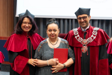 Three people stand next to each other during an academic ceremony. Two individuals wear red and black gowns and caps with decorative chains. The person in the center holds a closed red folder. In the background, a lecture hall with a screen and equipment is visible.
