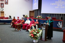 Six individuals in academic robes sit on stage, one individual stands at a podium speaking. In the background, a screen displays “100 years of the Faculty of Pharmacy WUM 1926–2026”. A floral arrangement stands in front.