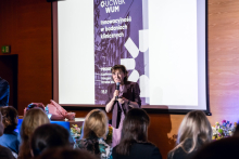  One person stands in front of a screen and speaks to the audience while holding a microphone. The screen displays the text “Innovation in clinical research UCWBK WUM.” Participants are seated in front of the stage.
