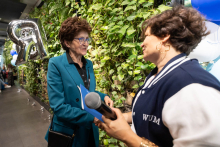 Two people stand facing each other near a green plant wall. The person on the left holds a blue folder and has a bag over the shoulder, while the person on the right holds a microphone. Silver balloons and a bright interior are visible in the background.