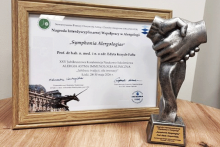 Dyplom w drewnianej ramie stoi na stole obok statuetki przedstawiającej splecione dłonie. Na dyplomie tekst z nagrodą „Symphonia Alergologia” oraz nazwiskiem odbiorcy. W tle widoczna grafika budynku.