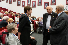 Several individuals sit in an auditorium. Three individuals stand and talk between rows of seats, with a wheelchair visible nearby. Portraits hang on the wall.