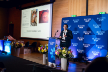 One person stands at a podium and delivers a presentation. Slides with charts and medical images are displayed on the screen. Flower arrangements and a UCWBK WUM logo backdrop are placed beside the stage.
