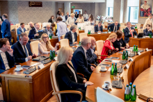 About 20 people are in a conference room. Some participants sit at tables, while others stand and move around. Microphones, documents, and drinks are placed on the tables.