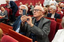 People sit on red seats in an auditorium. One person in the foreground holds a smartphone and takes a photo. Next to that person sits another attendee, while additional rows of participants are visible in the background, with hands resting on laps or armrests.