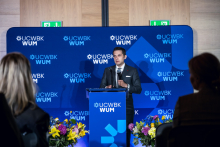 One person stands at a podium and speaks to the event participants. A UCWBK WUM logo backdrop is visible behind, with flower arrangements placed on both sides of the stage.