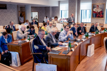 About 20 people are in a conference room. Some participants sit at tables, while others stand and move around. Microphones, documents, and drinks are placed on the tables.