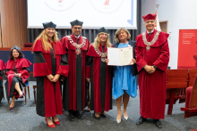 Five people stand in a lecture hall during a ceremony. Four individuals wear red and black academic gowns and caps, some with decorative chains. One person in a light blue dress stands in the center holding an open folder with a document. A presentation screen and wooden wall panels are visible in the background.