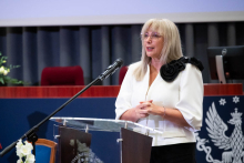 One person stands at a podium addressing the audience in a lecture hall. Elements of the stage and the university emblem are visible in the background.