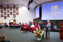 Seven individuals in academic robes sit on stage, one person stands at a podium and speaks. The screen displays “100 years of the Faculty of Pharmacy WUM 1926–2026”