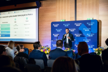 One person stands in front of the stage and speaks to the audience while holding a microphone. A presentation about the clinical research market in Poland is displayed on the screen.