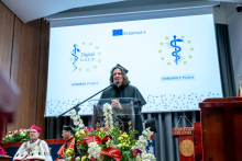 One person stands at a lectern and speaks into a microphone. A large screen behind displays Erasmus+ and project logos. Several officials sit in the background.