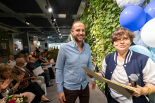 Two individuals stand next to each other in front of a green plant wall. The person on the right holds a microphone and documents, while the person on the left holds a sheet of paper. An audience is seated in the background, with some people holding documents.
