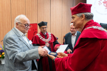 Two people stand close together during the presentation of an award. One person wears a light suit and holds an open case with a medal, while the other person in a red academic gown supports it while also holding the case. In the background, additional people in gowns are visible, including one holding a document, as well as a flower arrangement and wooden wall panels.