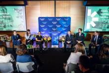 Six people sit on stage and take part in a panel discussion. One person holds a microphone and speaks, while the others sit beside.