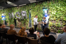 Two individuals stand in the middle of a room and exchange a document. The person on the right holds a microphone and a paper, while the person on the left receives the document. An audience sits on chairs in front of them, and a green plant wall with silver balloons forming the word “CSR” is visible in the background.