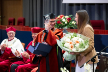 Seven individuals in academic robes sit on stage, one person stands at a podium and speaks. The screen displays “100 years of the Faculty of Pharmacy WUM 1926–2026”.