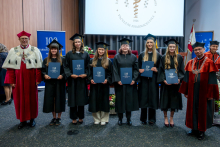 Seven graduates stand in a row holding folders during a ceremony. Two officials in academic robes stand on both sides. A university screen and banners are visible in the background.