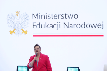 A woman, the Deputy Minister of Education, stands on stage and speaks into a microphone. In the background, the emblem and the text “Ministry of National Education” are visible.