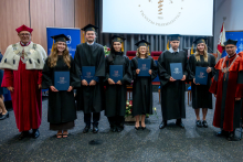 Six graduates stand in a row holding folders during a ceremony. Two officials in academic robes stand on both sides. The Medical University of Warsaw emblem is displayed on a screen behind them.