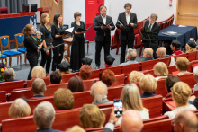 A group of performers dressed in black stands in a lecture hall holding open folders or sheet music and singing. On the left, one person holds a small child in their arms, and a child stands nearby without holding any objects. In the foreground, audience members sit in red seats, including one person holding a smartphone and recording or taking photos. A banner reading “Warszawski Uniwersytet Medyczny Wydział Lekarski” is visible in the background.