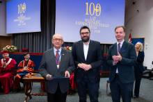 Three people stand on stage, one person holds a box and a document. In the background, individuals in academic robes are seated, and the screen displays “100 years of the Faculty of Pharmacy WUM 1926–2026”.