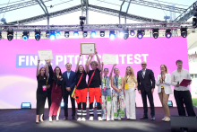 A group of several people stands on stage holding diplomas. In the background, a large screen displays the text “FINAL PITCH CONTEST.”