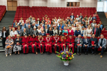 A large group poses for a group photo in an auditorium with red seats. In the front row, several people wear red and black academic gowns, some with decorative chains, and a few hold red folders. A large vase with colorful flowers is placed on the floor in front of them. Behind, more participants stand in rows, dressed in formal attire, some holding documents or folders. Additional rows of red seating and doors of the hall are visible in the background.