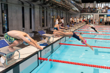 Women and men dive from starting blocks into a swimming pool during a competition. The participants wear swimsuits and are positioned above lanes marked with red dividers. Other people stand around and watch from the poolside and upper level.