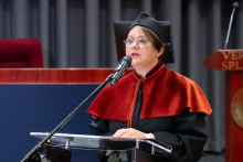 One person in an academic robe stands at a podium and speaks during a ceremony in a lecture hall. The stage is visible in the background.