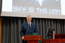 One person stands at a lectern marked with the inscription “VERITATIS SPLENDOR” and speaks into two microphones. On the lectern there are two bottles of water and a glass. Behind, a large screen displays the text “SKY IS THE LIMIT,” and stage elements including a monitor and seating are visible.