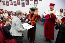 Two people in academic robes present a document and a medal to one person standing near the rows of seats. Event participants are seated around.