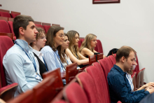 Several people sit in rows of red auditorium seats and face forward, listening attentively. Some wear lanyards and hold notebooks or devices. In the foreground, one person uses a laptop. The background shows additional rows of seating and a lecture hall interior.