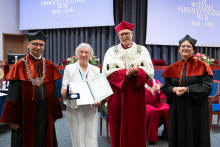 Four people stand on stage, one person holds an open document and a medal. In the background, a screen displays “100 years of the Faculty of Pharmacy WUM 1926–2026”.