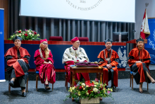 Five people sit on a stage during an academic ceremony. A central table stands in front of them. Flower arrangements are placed near the stage. In the background, a projection screen and a university emblem are visible. A national flag stands to the right.