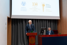 One person stands at a lectern with the inscription “VERITATIS SPLENDOR” and speaks into microphones. Two bottles of water and a glass are placed on the lectern. Behind, a large screen displays the logos of the Medical University of Warsaw and the Faculty of Pharmacy, along with the text “100 lat Wydziału Farmaceutycznego WUM 1920–2020.” Another person sits at a desk to the side. The background features a large projection screen and vertical wall panels.