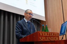 One person stands at the same lectern labeled “VERITATIS SPLENDOR” and speaks into microphones. Two green bottles of water and a glass are placed on the lectern. A computer monitor is positioned to the side. In the background, part of a large projection screen with university logos and text related to the Faculty of Pharmacy is visible.