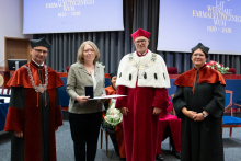 Four people stand on stage. One person holds a document and a case. Behind them, a screen shows “Faculty of Pharmacy WUM 1926–2026