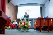 One person stands at a lectern and speaks. One person sits behind on the stage. A large flower arrangement stands in front of the lectern. A projection screen with a university emblem is displayed in the background.