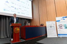 One person stands at the lectern marked “VERITATIS SPLENDOR” and speaks into microphones, while another person sits behind at a desk. On the right, two standing banners display information and equipment graphics, including the logo “CEMIS TECH” and images of laboratory devices. Above, a large screen shows multiple sponsor logos and text. The setting is a lecture hall with wooden wall panels and stage equipment.