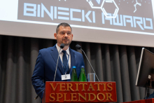 One person stands at a lectern with the inscription “VERITATIS SPLENDOR” and speaks into two microphones. Two green bottles of water and a glass stand on the lectern. Behind, a large screen displays the word “BINECKI” along with a scientific-style graphic.