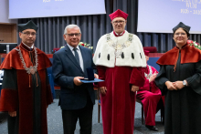  Four people stand on stage. One person holds a document and a case. Behind them, a screen displays “Faculty of Pharmacy WUM 1926–2026”.