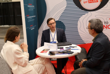 Three individuals sit at a round table and engage in a conversation during a trade fair event. Brochures, phones, notebooks, and a card reading “Medical University of Warsaw” are placed on the table. Elements of the exhibition stand with informational text and graphics are visible in the background.