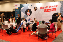 n an exhibition space, several groups of individuals sit at tables and hold discussions. Chairs and tables with promotional materials are arranged on a red carpet. A large wall in the background displays the text “Study and Research in Poland!” along with graphics related to international education.