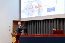One person stands at a lectern marked “VERITATIS SPLENDOR” and speaks into microphones while looking toward a large screen. The screen shows a diagram of a synapse with labeled elements including “AChE/BuChE inhibitors.” Another person sits behind at a desk with a laptop.