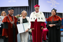  Four people stand on stage. One person holds a document and a case. Behind them, a screen displays “Faculty of Pharmacy WUM 1926–2026”.